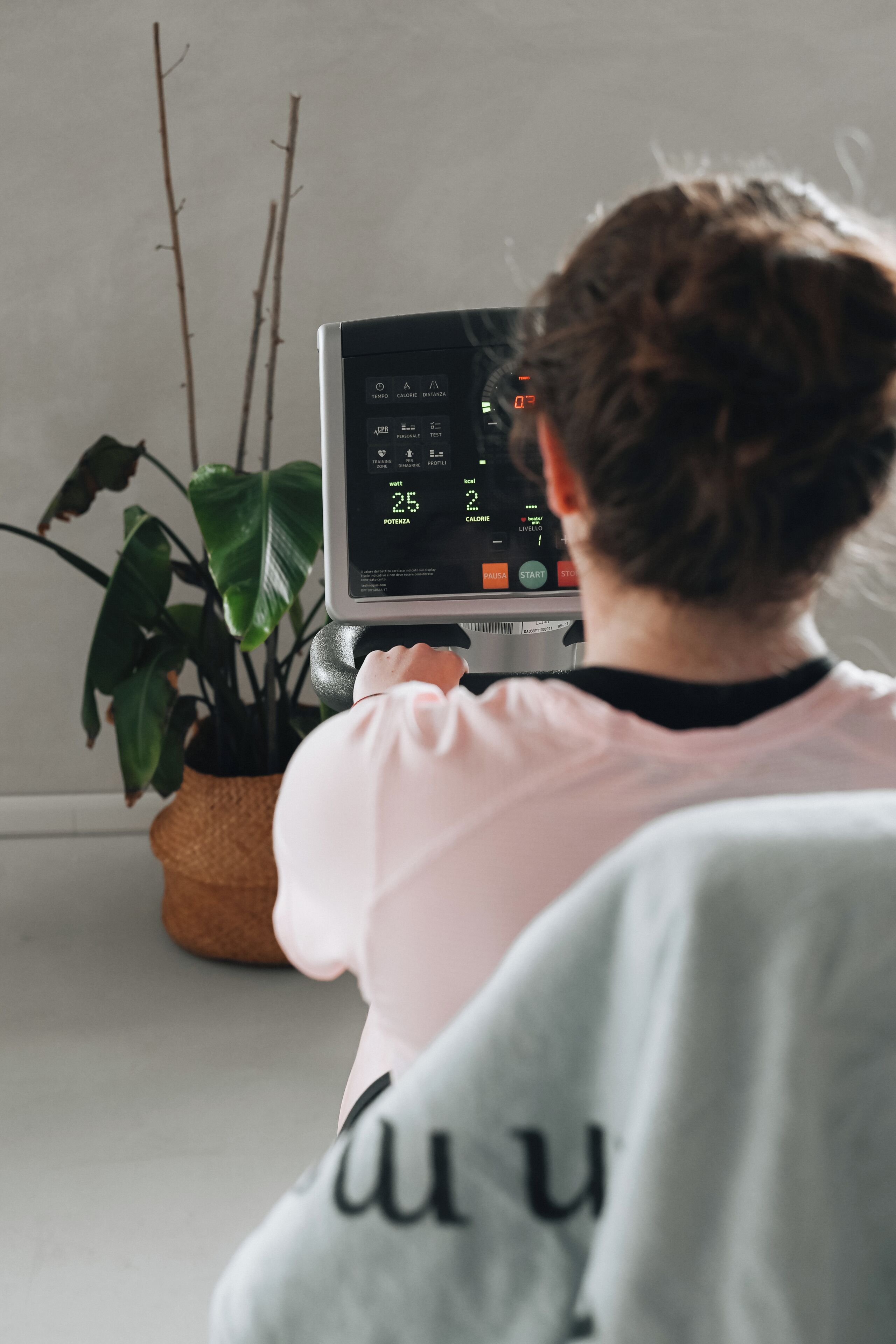 A person using a stationary bike with a digital display, a potted plant, and a wall in the background.