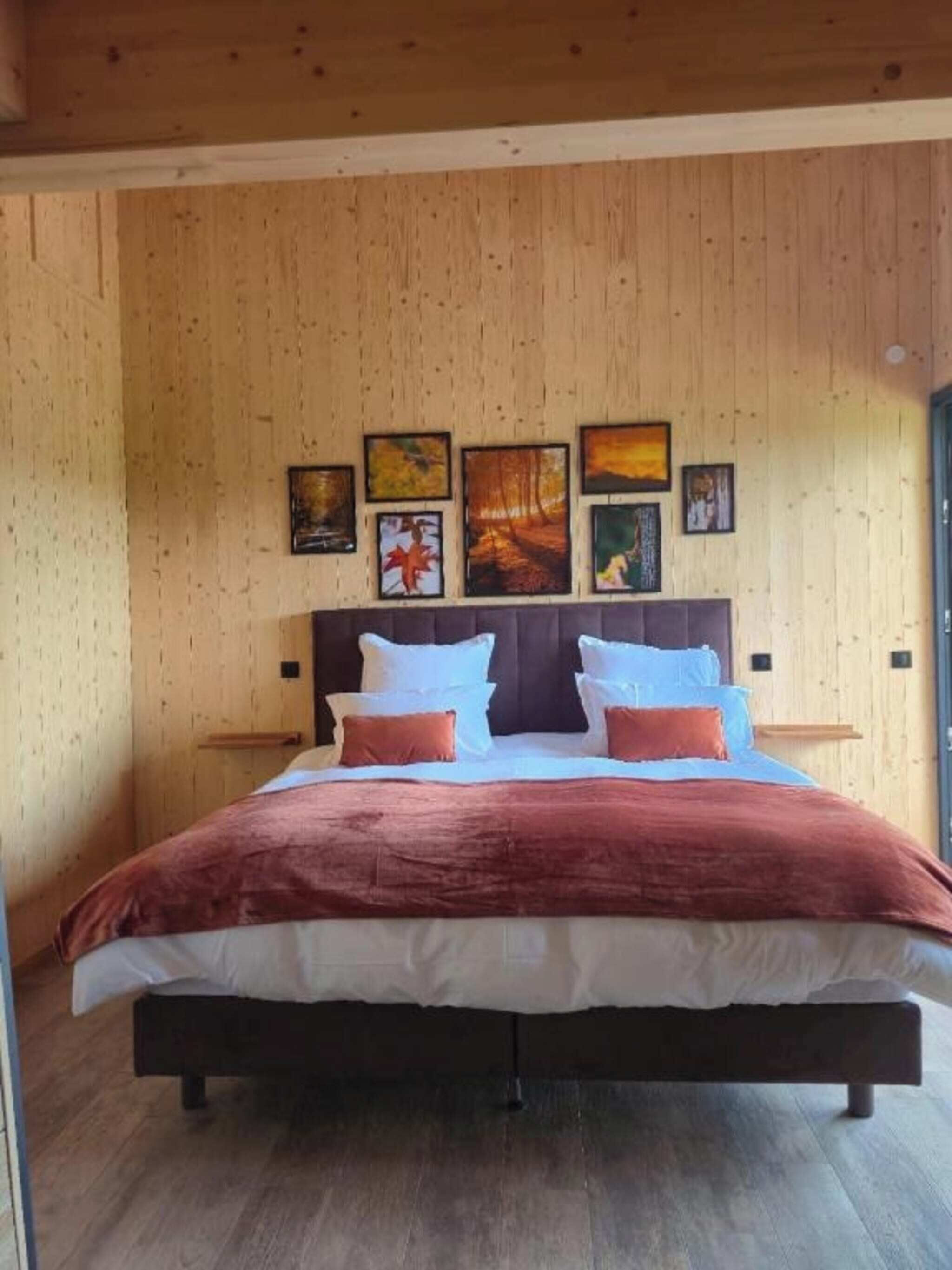 A bedroom with a wooden wall, a bed with a brown bedspread, and several framed pictures on the wall.