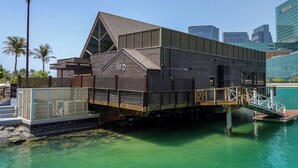 Un edifici a la riba del mar amb un teulat inclinat, una terrassa de fusta i una escala que baixa a un moll.