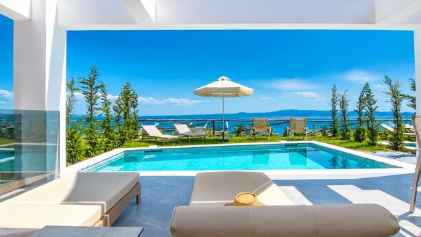 A modern outdoor pool area with lounge chairs and an umbrella, overlooking a clear blue sky and a distant landscape. - Sunrise Villas Olea Collection (Chaniotis)