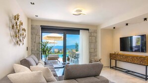 A modern living room with a grey sofa, a flat-screen TV, and a view of a pool and mountains. - Sunrise Villas Olea Collection (Chaniotis)