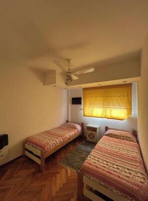 Una habitación pequeña con dos camas individuales, un ventilador de techo, una ventana con cortinas amarillas y piso de madera.