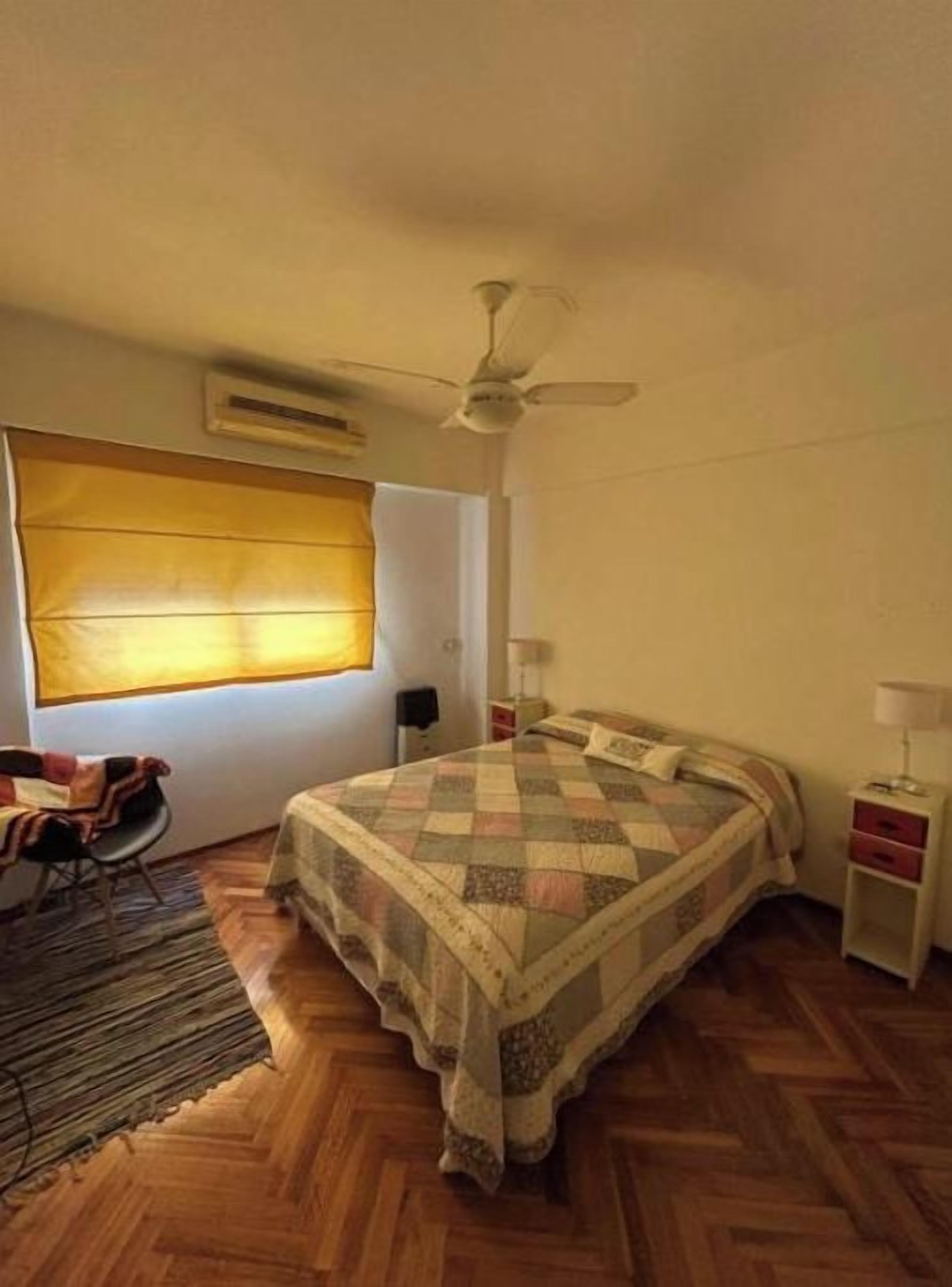 Un dormitorio con cama, ventilador de techo, ventana con persianas, mesita de noche y una silla.