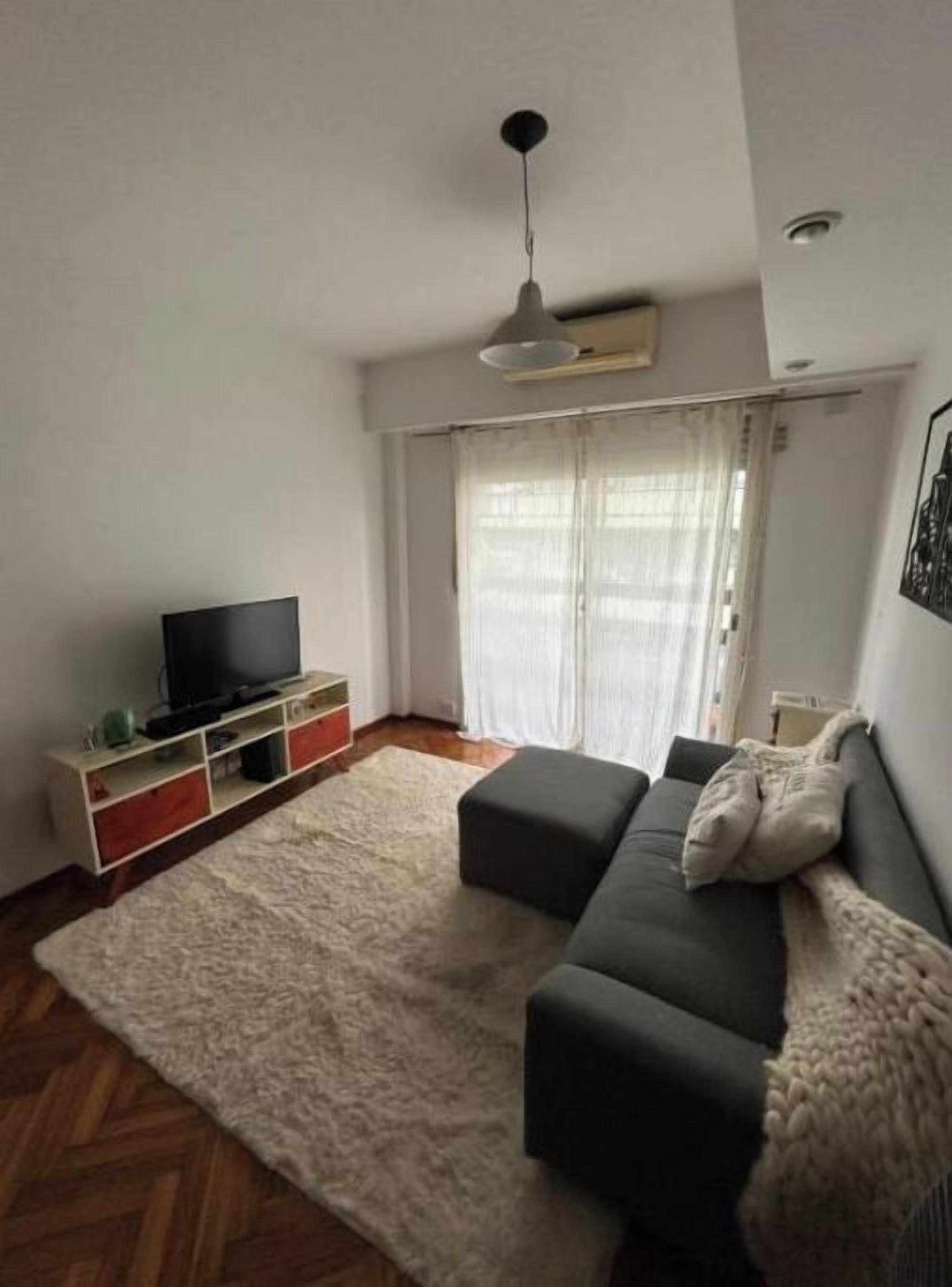 Een woonkamer met een bank, een televisie, een wit vloerkleed en een raam met gordijnen.