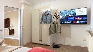 Ein Schlafzimmer mit einem Bett, einem roten Kopfkissen, einem Kleiderschrank und einem Fernseher, der einen Sportkanal zeigt.