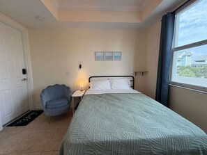 A bedroom with a bed, a chair, a nightstand, a lamp, and a window with a view of houses. - Relaxed Studio – Beach Gear – Near Barefoot (Bonita Springs)