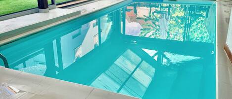 Piscina interior com paredes de vidro transparente, chão em azulejo e estrutura de teto em madeira.