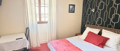 A bedroom with a bed, a small table, a chair, a window with curtains, and a wall with a geometric pattern.