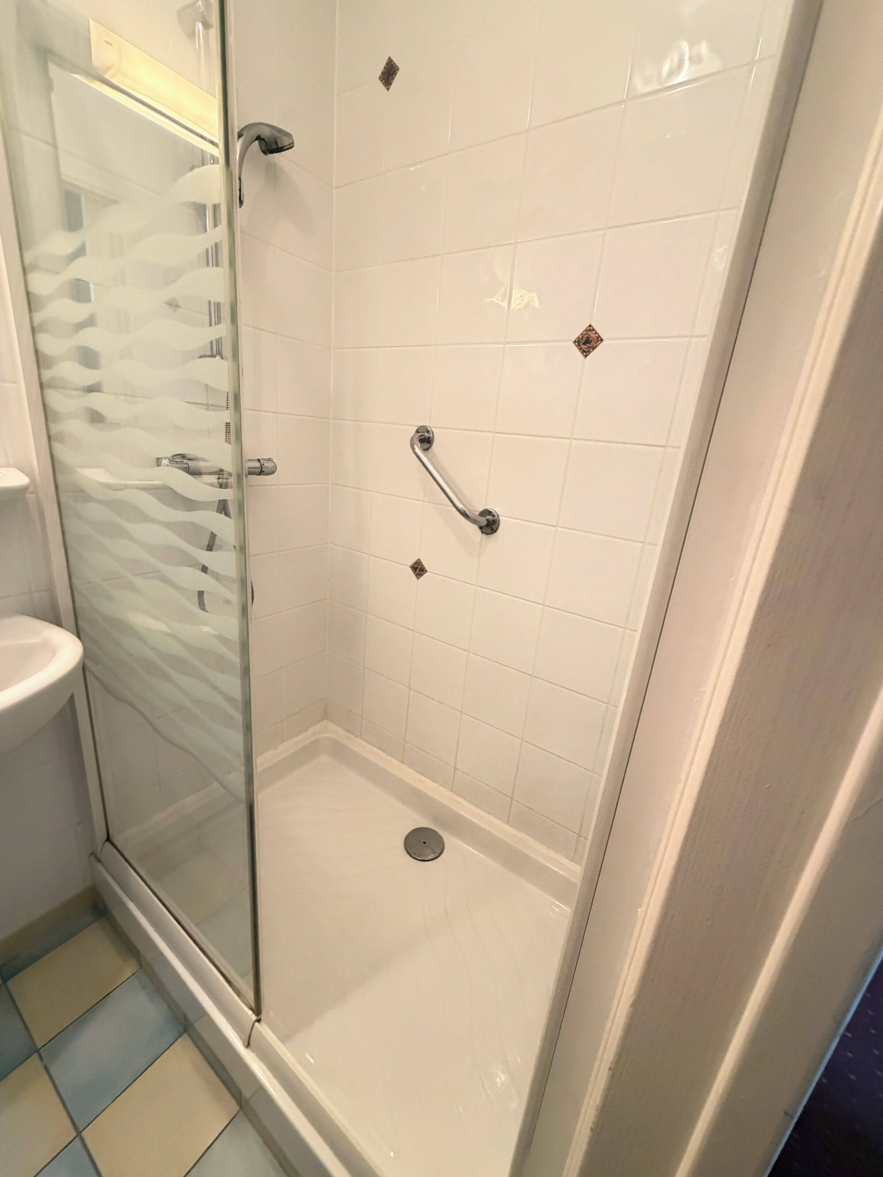 Une douche fermée par des parois de verre, avec des carreaux blancs, une pomme de douche à main et un système de commande de douche à main.