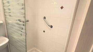 Une douche fermée par des parois de verre, avec des carreaux blancs, une pomme de douche à main et un système de commande de douche à main.