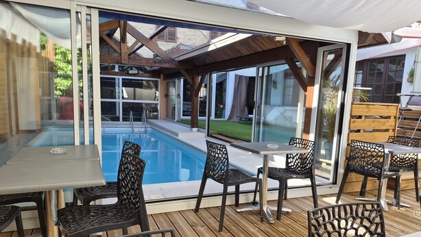 An outdoor pool area with glass walls, wooden deck, and metal chairs. - HOTEL DE PARIS  (Poligny)