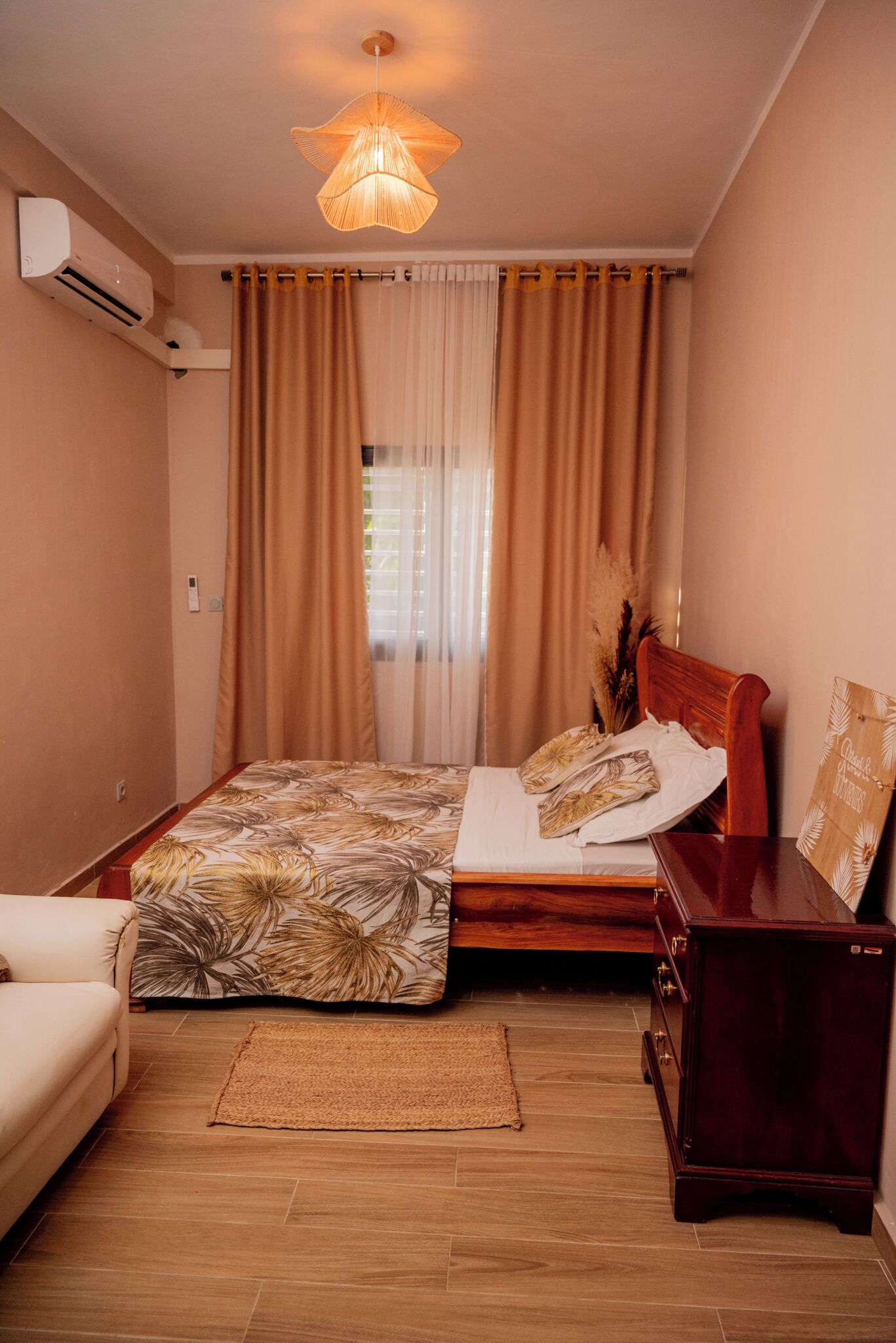 Um quarto com uma cama, uma mesa de cabeceira de madeira, uma cadeira, um penteou de madeira, uma luminária no teto e uma janela com cortinas.