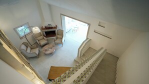A spacious living area with a high ceiling, a dining table, two armchairs, and a sofa. A staircase with patterned railings leads to an outdoor area.