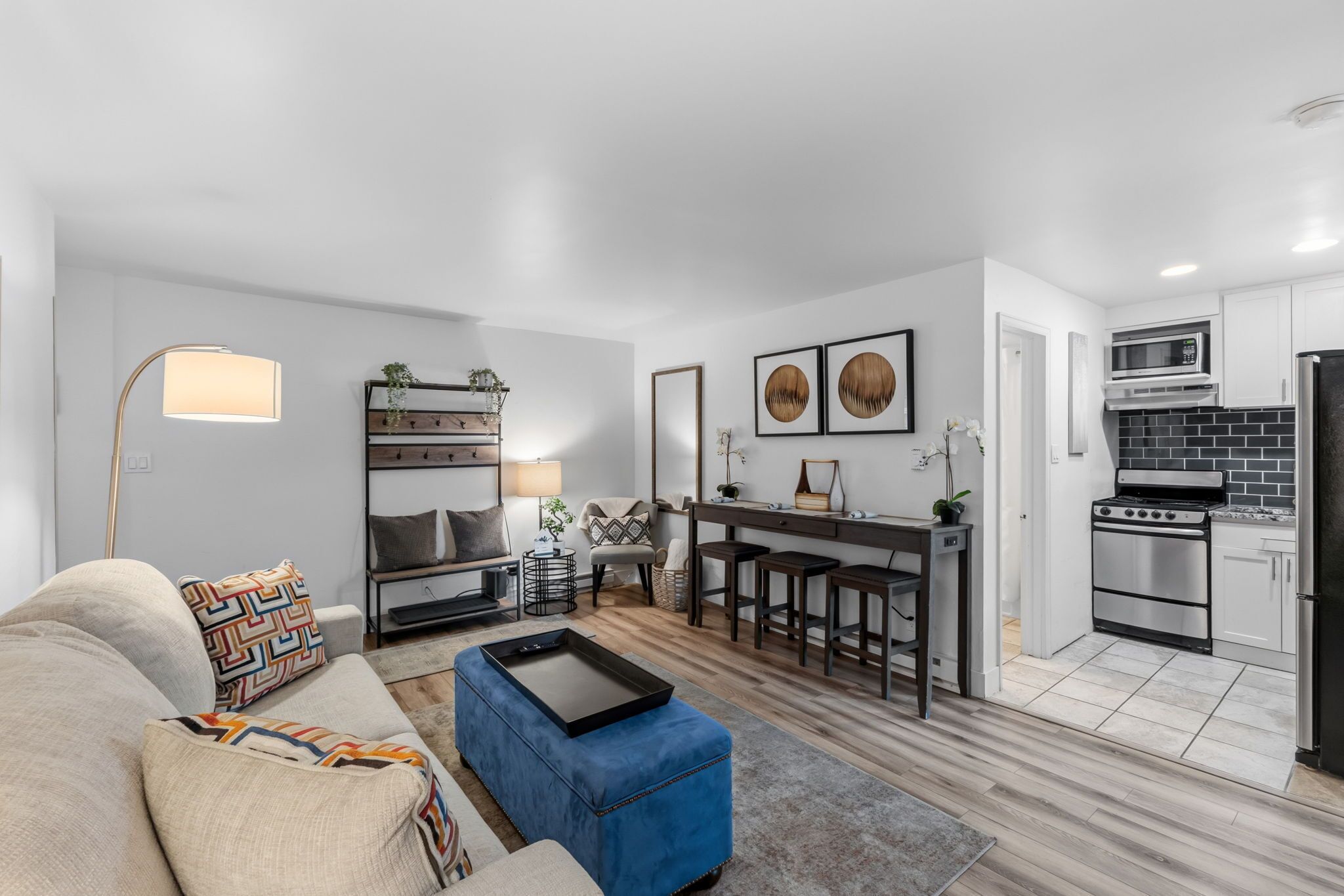 A modern living room with a sofa, ottoman, and a coffee table. Adjacent is a kitchen with a microwave, stove, and bar stools.