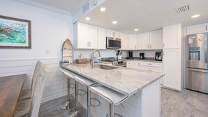 A modern kitchen with white cabinetry, a marble countertop, stainless steel appliances, and a bar stool area. - Cinco Brisas 2 3 Bedroom Home (Orange Beach)