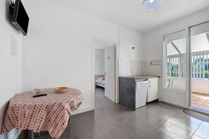 Ein Zimmer mit einem Bett, einem kleinen Tisch mit gemustertem Tischläufer, einem an der Wand befestigten Fernseher und einer Küchenzeile mit Geschirrspüler.