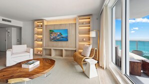 Een moderne woonkamer met een grote televisie, witte banken, een rieten fauteuil en een houten salontafel.