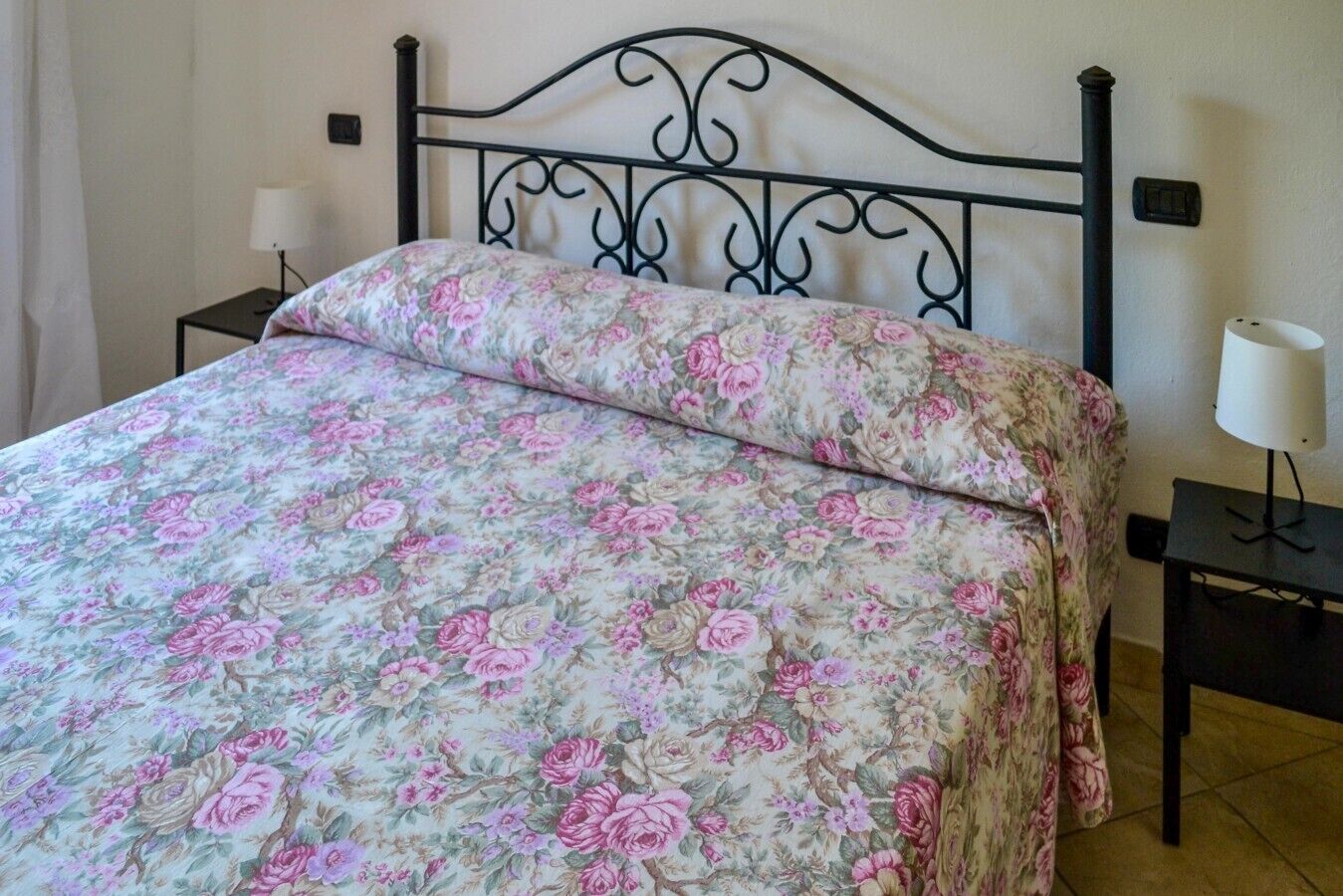 A bed with a floral bedspread, a black metal headboard, two bedside tables with lamps, and a plain wall.