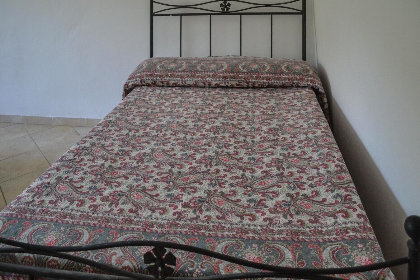A single bed with a patterned bedspread in a room with a plain wall and a tiled floor.