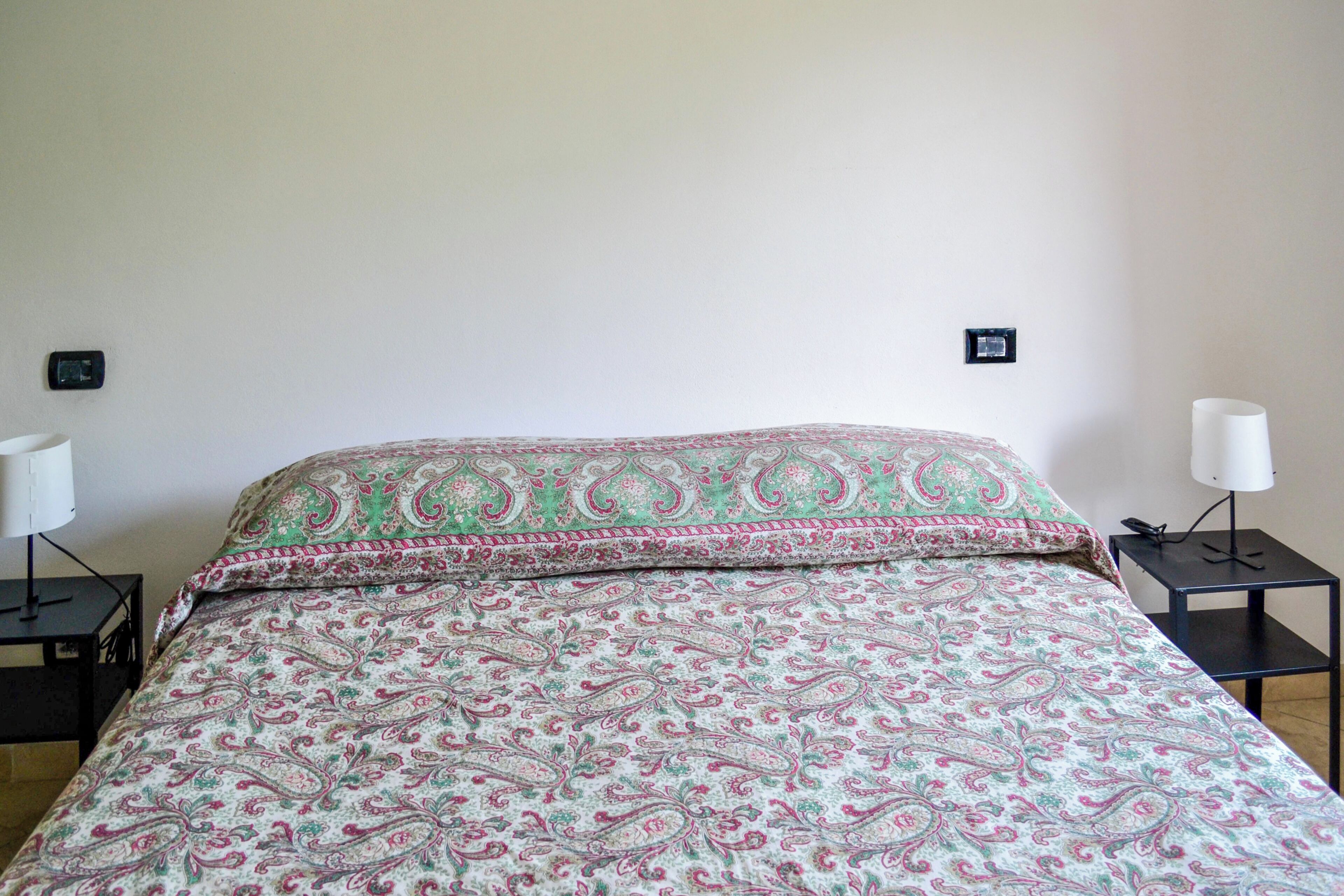 A bed with a patterned bedspread, two bedside tables with lamps, and wall-mounted switches.