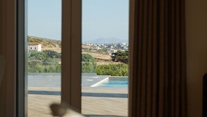 A room with a large window, a view of a swimming pool, and a glimpse of the surrounding landscape. - Andros Royal Villa (Koumari)