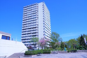 Ein hohes, modernes Gebäude mit mehreren Balkonen, umgeben von einer gepflegten Grünanlage mit Bänken und Bäumen.