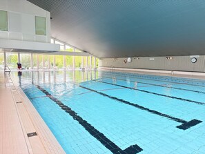 Ein Hallenbad mit deutlich markierten Bahnen, einer gefliesten Liegefläche und einem überdachten Bereich mit Liegen und einem Rettungsschwimmerstuhl.
