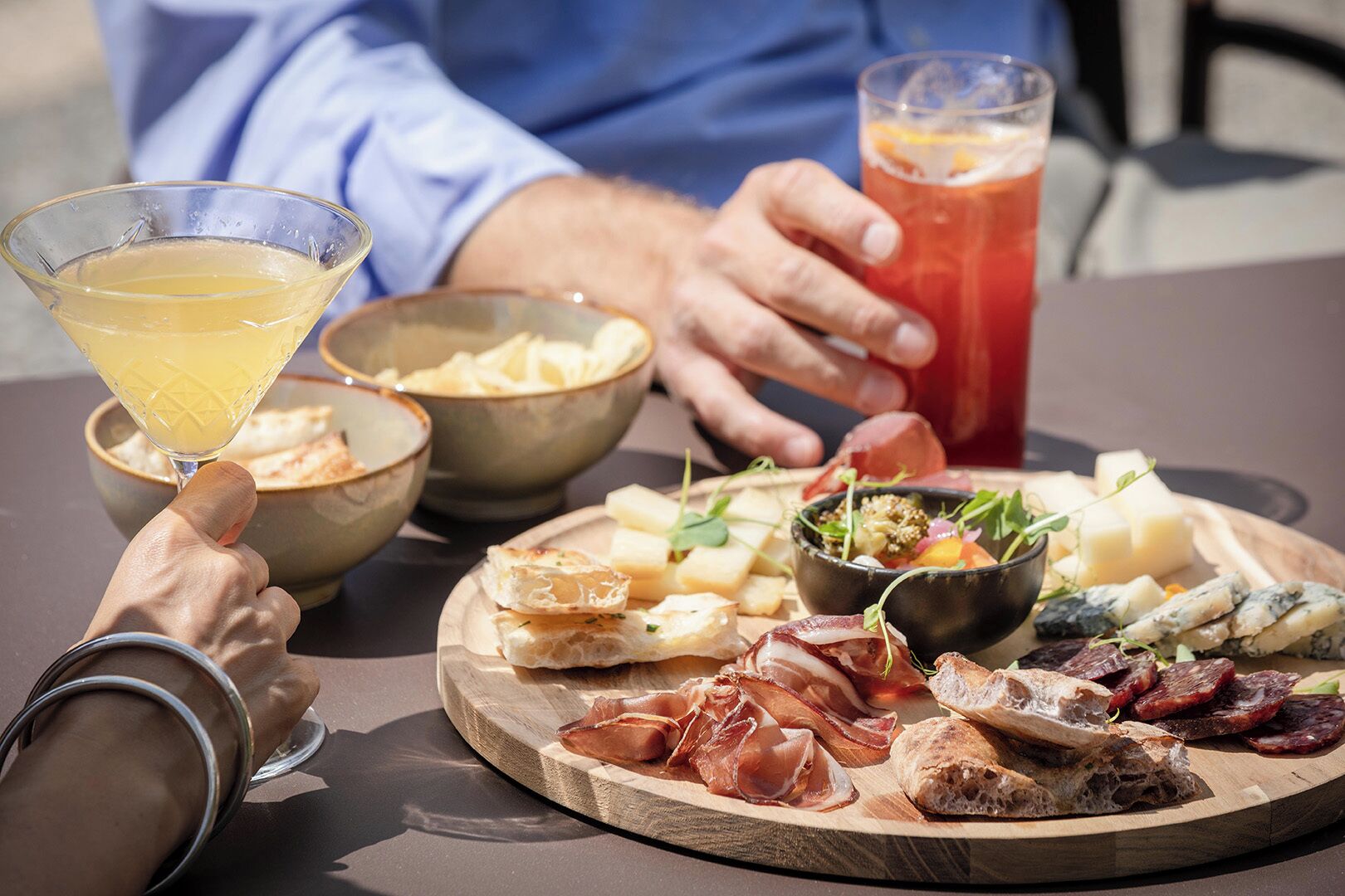 Une table garnie d’une variété d’aliments, notamment du fromage, de la charcuterie et du pain, ainsi qu’un verre à martini et un verre d’une boisson rouge.