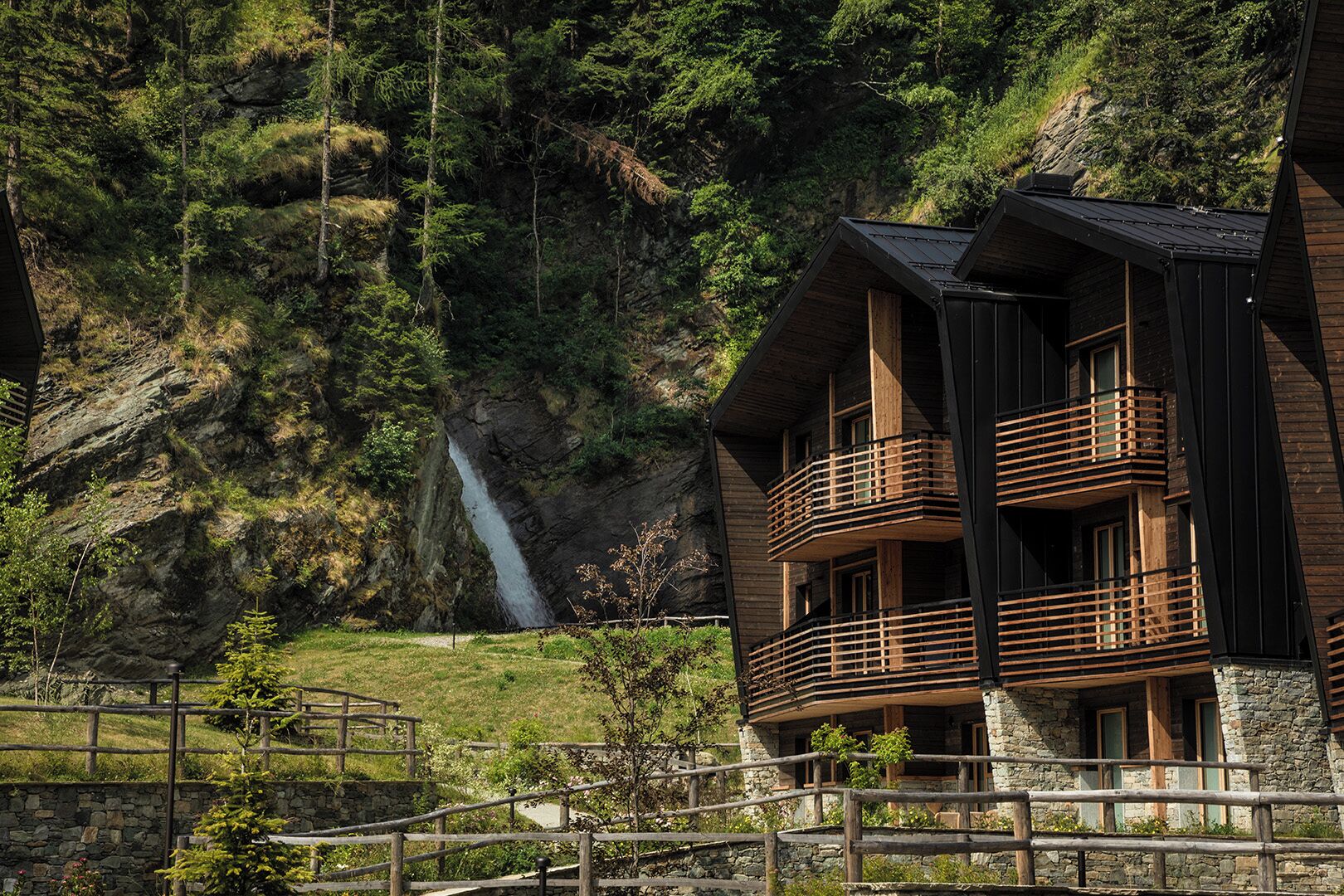 Un immeuble en bois à plusieurs étages, doté de balcons, qui se dresse sur fond de falaise rocheuse et de cascade.