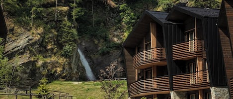 A multi-story wooden building with balconies, set against a backdrop of a rocky cliff and a waterfall.