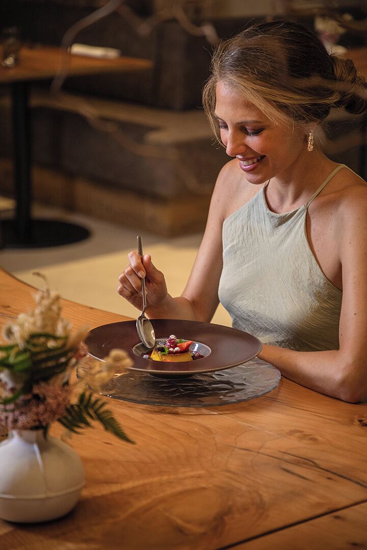 Iemand zit aan een houten tafel met een bord eten, een lepel en een vaas bloemen.