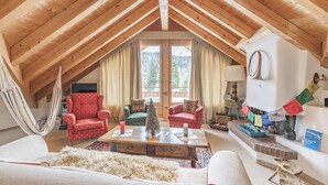 A cozy loft-style living room with a high wooden ceiling, a hammock, and a fireplace with colorful decorations. - Ski-in · Luxury Alpine Apartment · Celerina (Celerina/Schlarigna)