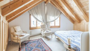 A bedroom with a sloped ceiling, a bed, a chair, a small table, and a window with curtains. - Ski-in · Luxury Alpine Apartment · Celerina (Celerina/Schlarigna)