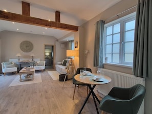 Ein modernes Wohnzimmer mit Essbereich, Holzbalken, einem runden Tisch und einem großen Fenster mit Vorhängen.