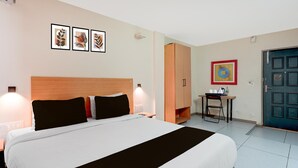 Et hotelværelse med en seng, sengeborde med lamper, et skrivebord med stol, et garderobeskab og billeder hængende på væggen.