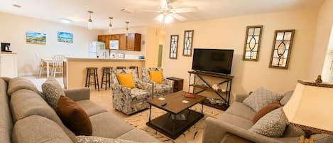A living room with a sofa, armchairs, a coffee table, and a TV. There is a kitchen area in the background.