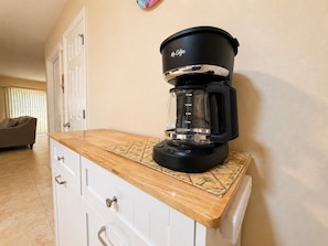 Une machine à café posée sur un plan de travail dans la cuisine, avec un tapis à motifs, un meuble blanc avec des tiroirs et une porte visible au fond.