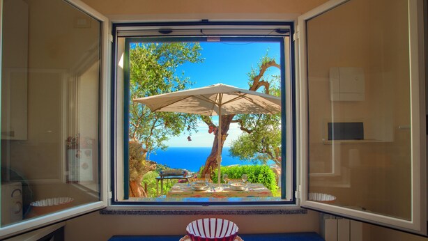 Una habitación con ventana que da a un comedor con sombrilla, una mesa y vistas al mar.