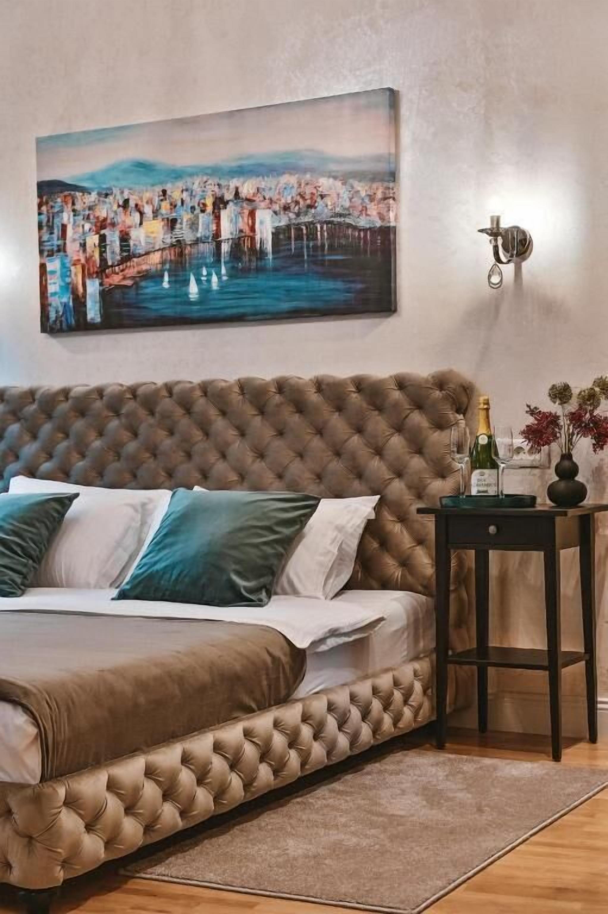Un dormitorio con cabecera capitonada, cama con almohadas blancas y azul turquesa, un cuadro de un paisaje urbano, una mesita de noche con una botella y un florero con flores.