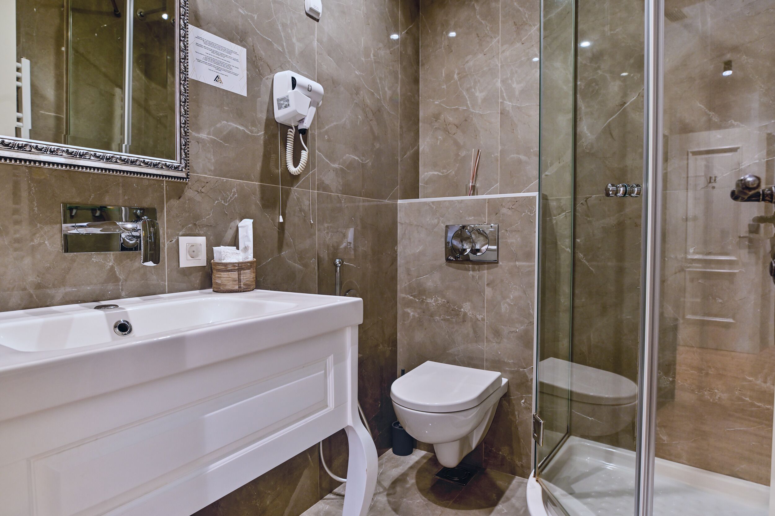 Baño con lavamanos blanco, inodoro, ducha con cerramiento de vidrio y secador de pelo montado en la pared.