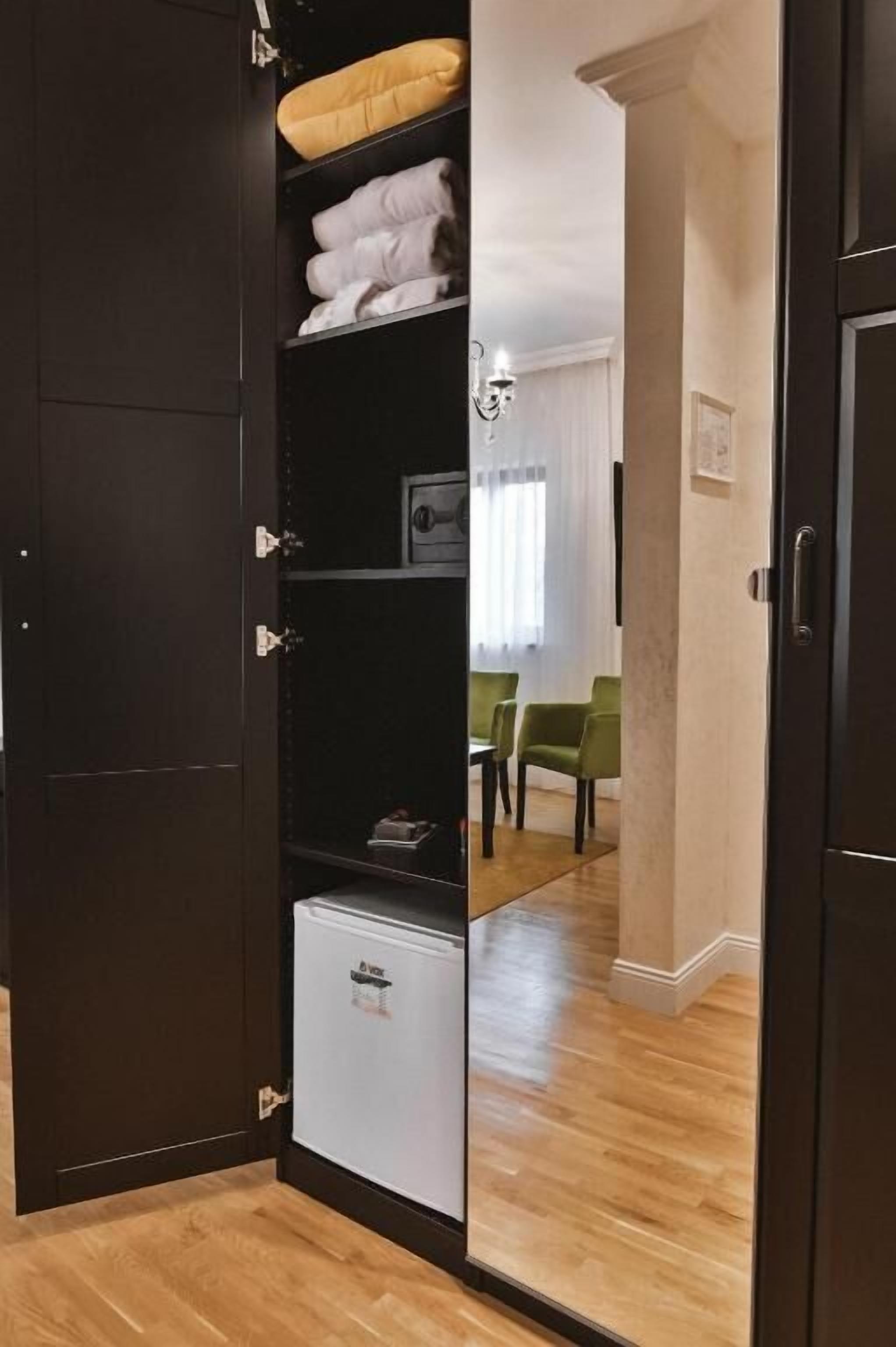 A room with a wardrobe, a mirror, a small fridge, and a green chair.