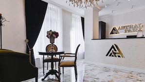Un área de recepción con piso de mármol, una mesa de recepción con el cartel 'Astral Park', una araña y una mesa con sillas.