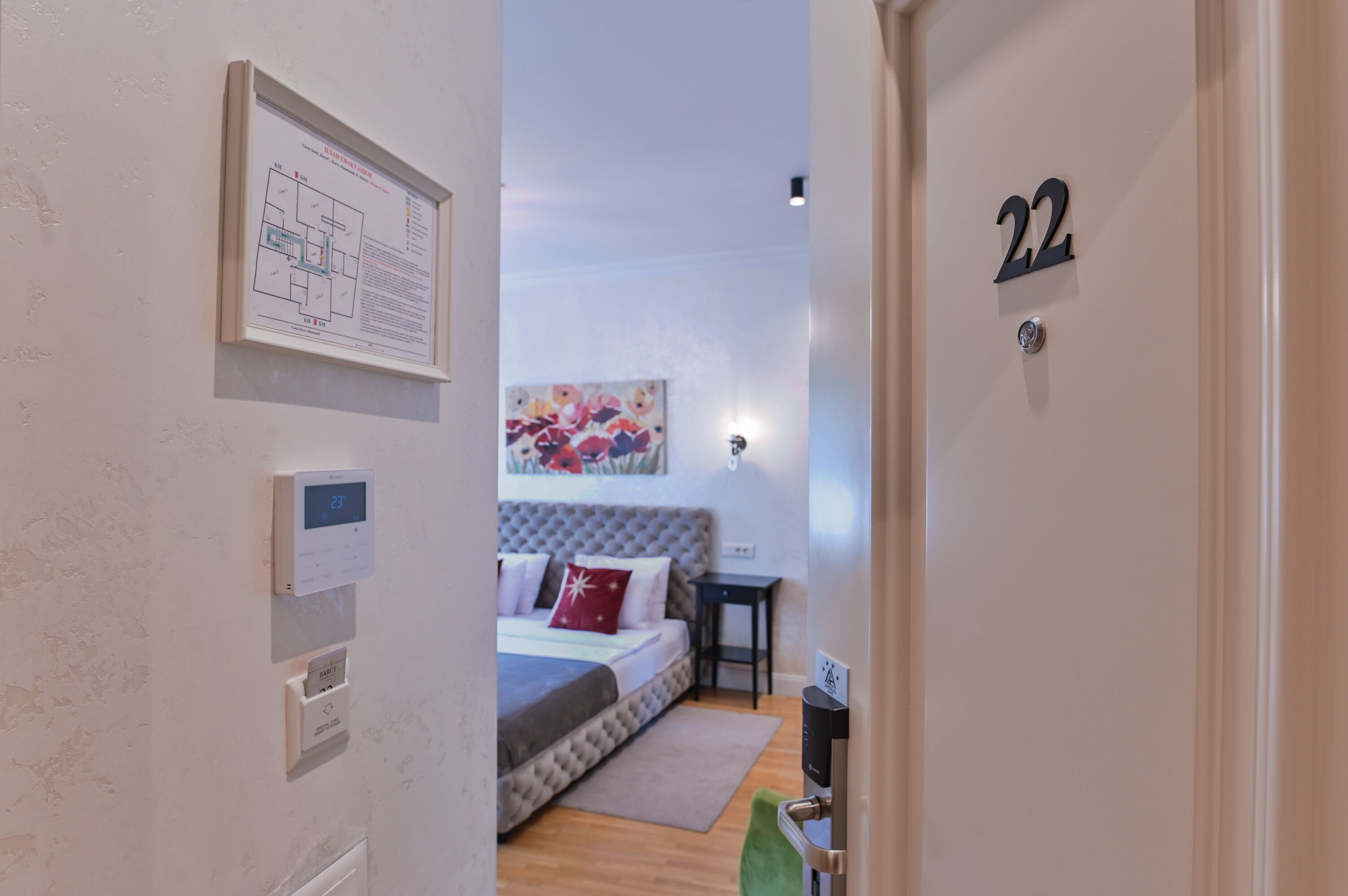 Habitación de hotel con cama, una mesita, termostato fijado a la pared, mapa enmarcado y una puerta con el número 22.