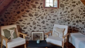Ein gemütliches Dachzimmer mit Steinwänden, Holzbalken, zwei weißen Sesseln, einem kleinen Tisch und einem Fenster.