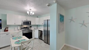 A kitchen with white cabinets, a stainless steel refrigerator, a microwave, and a dishwasher. There is a dining table with chairs and a decorative mat. - Estero Beach & Tennis 303B | Beachfront Studio + Resort Amenities (Fort Myers Beach)