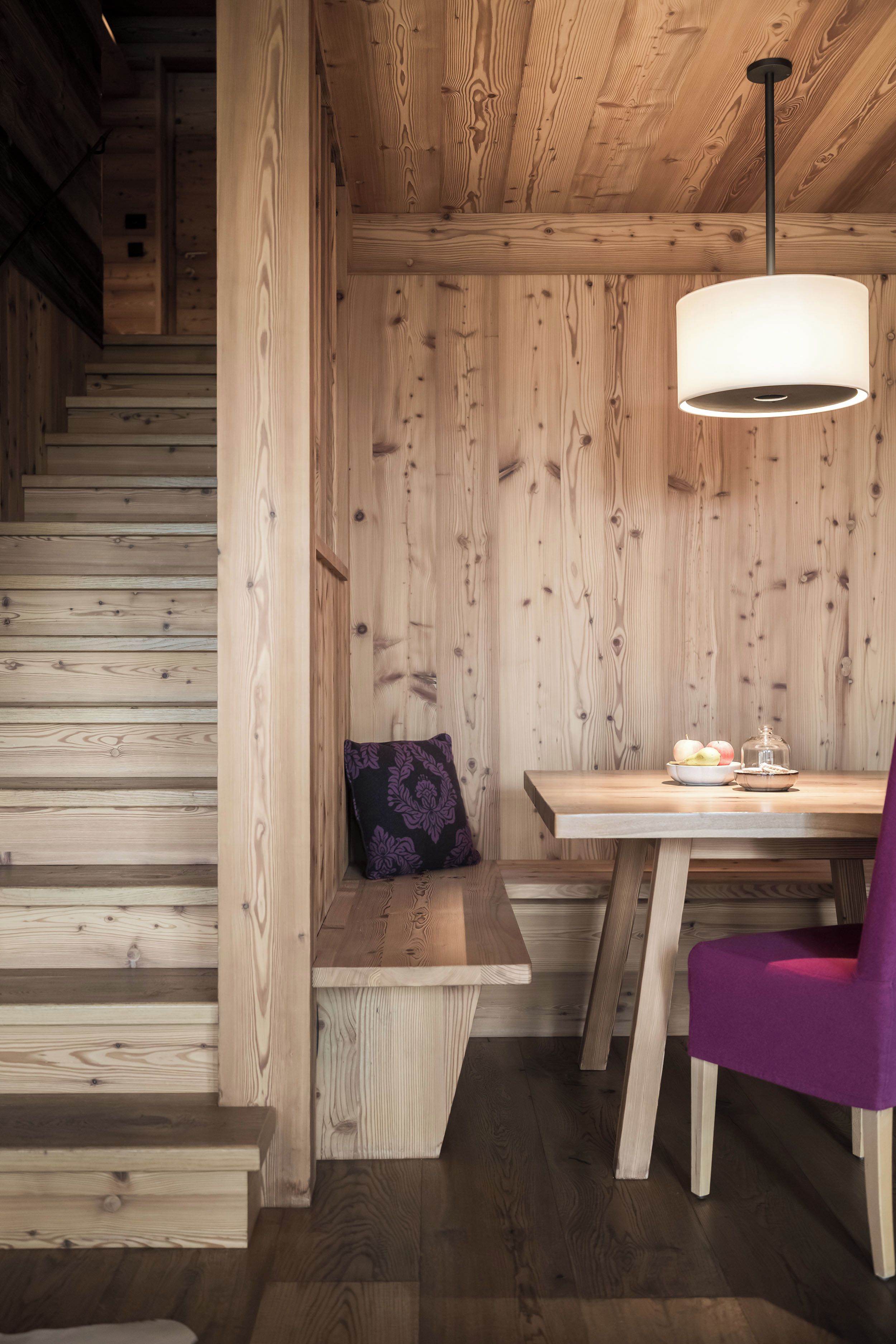 Un interior de madera con una escalera, una mesa con lámpara y una silla morada.