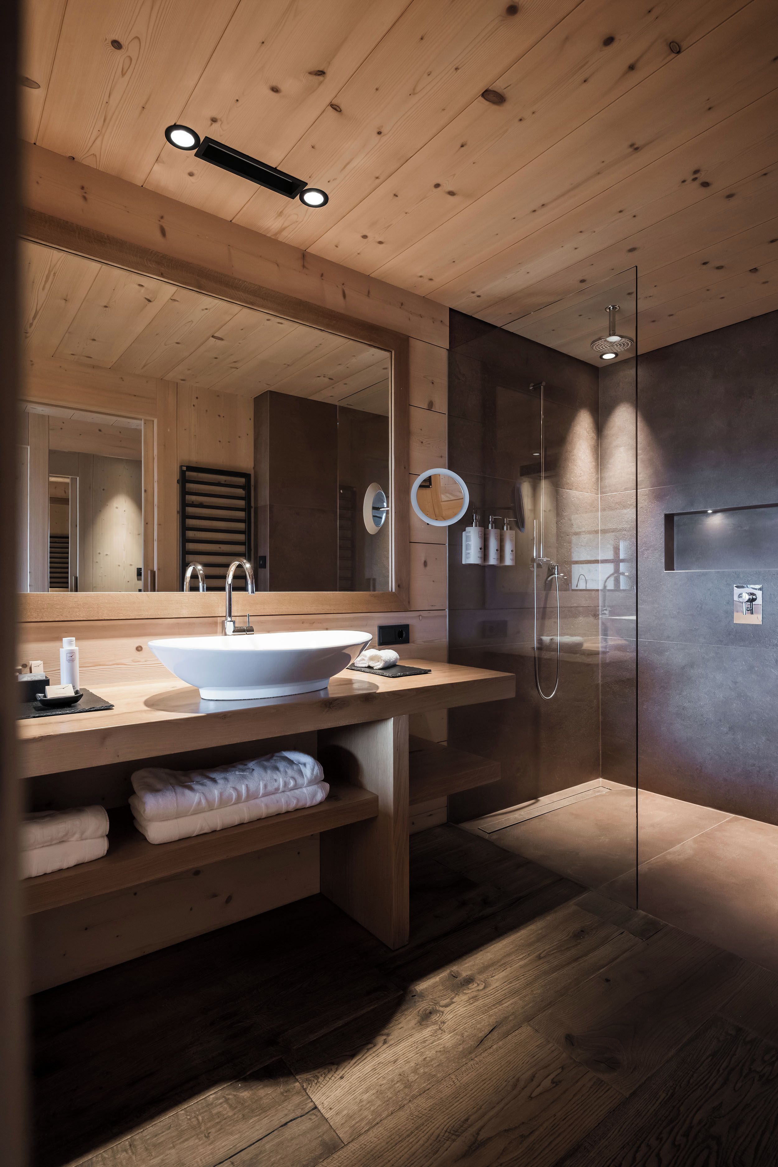 Un baño moderno con techo de madera, un espejo grande, un lavamanos, ducha y zona de ducha con mampara de vidrio.