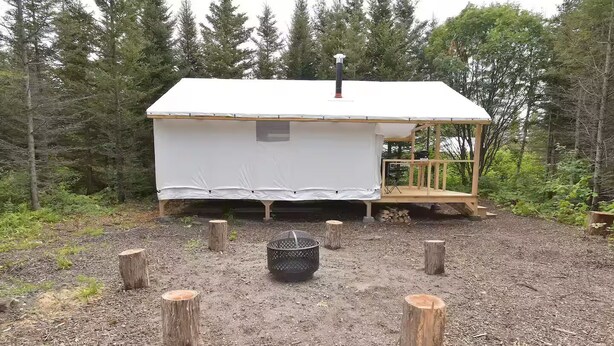 Eine kleine Holzhütte mit weißer Fassade, einer Terrasse und einem Feuerkorb, der von Holzstümpfen umgeben ist.