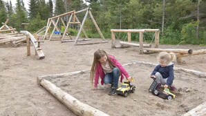 Zwei Kinder spielen mit Spielzeugautos in einem Sandspielplatz mit Holzkonstruktionen im Hintergrund.