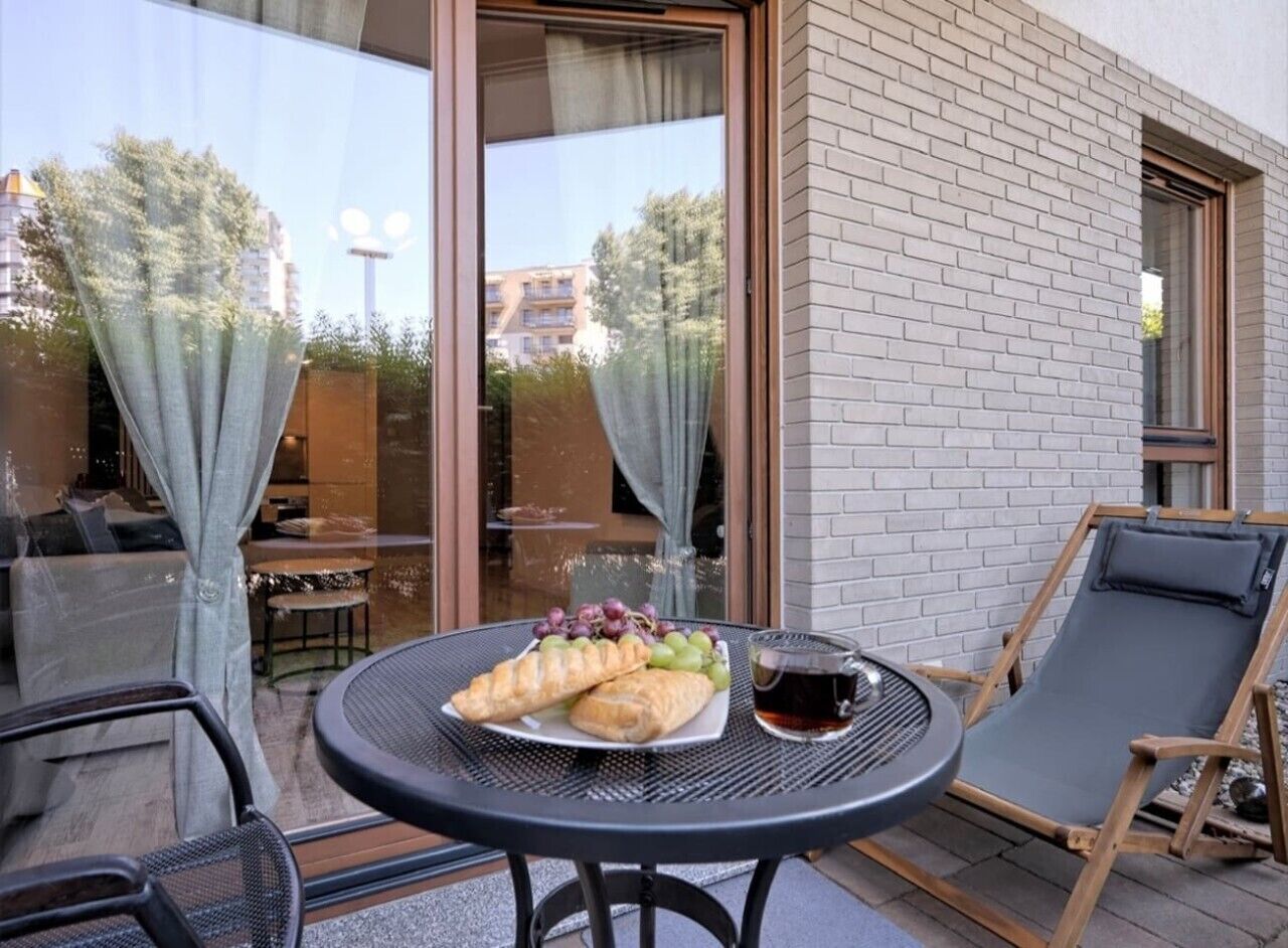 A table with croissants and grapes, a chair, and a glass of beverage outside a building with a brick wall and large windows.